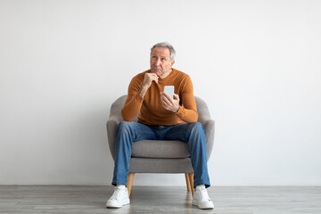Portrait of mature man using smartphone sitting on armchair - obrazy, fototapety, plakaty
