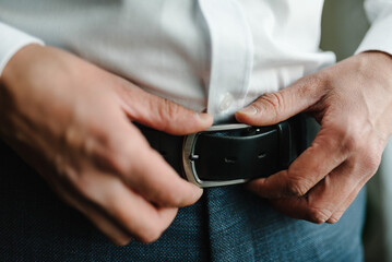 A man in pants and shirt buttoned a black leather trouser belt. Groom dress up a belt with a...