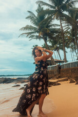 vacation leisure. young brunette girl in black stylish dress is standing in profile with hands near hair and looking away on a beach coast background near ocean. travel lifestyle concept, free space