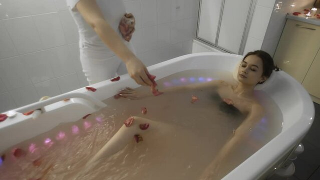 A dark-haired girl is lying in a Jacuzzi bath. A female employee at a spa salon pours rose petals into a Jacuzzi tub. The concept of a healthy lifestyle and body care.
