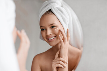 Portrait of young lady applying nourishing eye cream after shower, looking at mirror and smiling in...