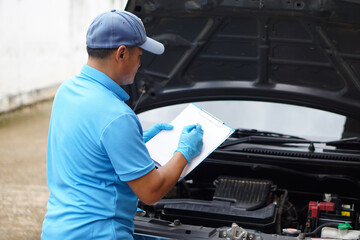 Asian mechanic is writing something on paper clipboard during checking car engine. Concept : Car maintenance service at home.