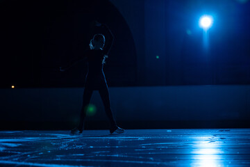 Young female figure skater is performing woman's single skating choreography on ice rink. Dark silhouette of woman practicing skills on ice arena against background of soft blue light and spotlights. © kinomaster