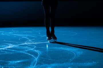 Dark silhouette of female legs in figure skating skates sliding on ice arena. Young woman trains on...