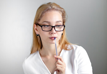Thinking caucasian young woman in white and wearing glasses holding pen on lips on gray background