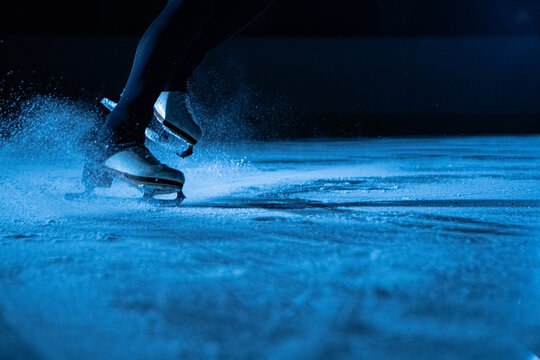 Detailed shot of women's legs in white figure skating skates on cold ice arena in the dark with blue light. A woman slides on the ice, splashing particles of sparkling ice into the camera. Close up.