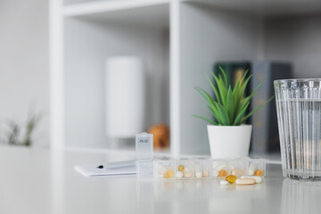 Closeup of medical pill box with doses of tablets for daily take a medicine for treatment, cure the disease. Glass cup of water on table, gray background. Green flowerpot or plant. Pen and notepad