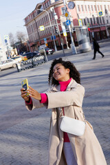 Happy curly brunette girl doing selfie on phone outdoors. Young woman happy walking in street. Pink sweater, beige coat, beige hat. Sun in city. Fashionable asian girl with frizzly hair with iphone