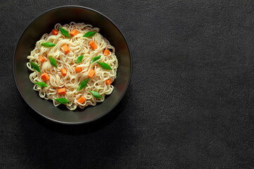 Noodles instant cooked with vegetables carrot and green onion dark bowl, on dark background, top view, space to copy text.