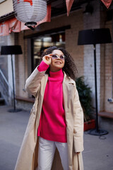 Happy curly brunette girl in sun glasses and hat smiling outdoors. Young woman happy walking in street. Pink sweater, beige coat, beige hat. Sun in city. Fashionable asian girl with frizzly hair