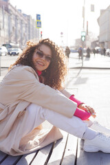 Happy curly brunette girl in sun glasses and hat smiling outdoors. Young woman happy walking in street. Pink sweater, beige coat, beige hat. Sun in city. Fashionable asian girl with frizzly hair