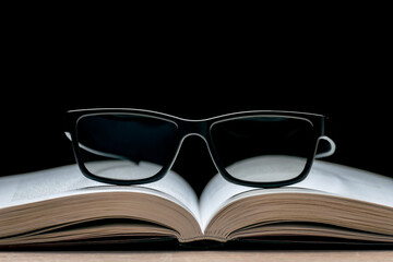 Eyeglasses lie on an open book on a black background. Reading Glasses. Classic black frame glasses.