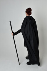 Full length portrait of pretty redhead female model wearing black futuristic scifi leather costume, black cloak holding a spear weapon. Dynamic standing pose on white studio background.