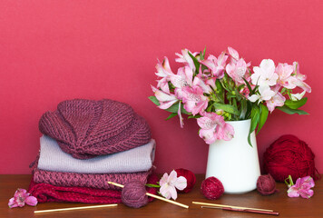 Hand crocheted burgundy beret and scarf. A set of crochet hooks, balls of wool yarn and a bouquet of fresh alstroemeria flowers on a wooden table against a red background. Spring needlework concept