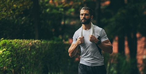 Determined young man running outside in the park. Fit boy doing exercise outdoor. Beautiful sporty male run alone. Sweaty athlete training with towel. Wellness, health, sport, fitness open air concept