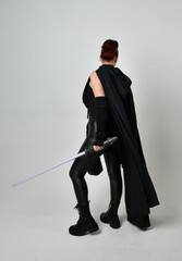 Full length portrait of pretty redhead female model wearing black futuristic scifi leather cloak costume, holding a lightsaber weapon. Dynamic standing pose on white studio background.