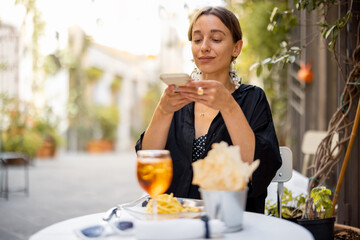 Young woman takes photo on phone of pasta and wine, having lunch at restaurant on cozy street in old italian town. Concept of italian cuisine and lifestyle