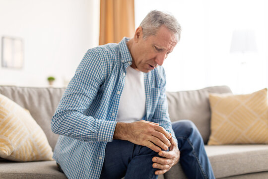 Mature man with knee pain sitting on couch at home - Powered by Adobe