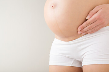 Young adult pregnant woman with naked big belly isolated on light gray background. Hand touching skin with caesarean scar before second baby birth. Closeup. Side view.