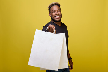 A positive strong black guy with a smile on his face holds two white bags of food in one hand. A friendly athletic man has picked up his bags, invites you to shop at a major shopping center