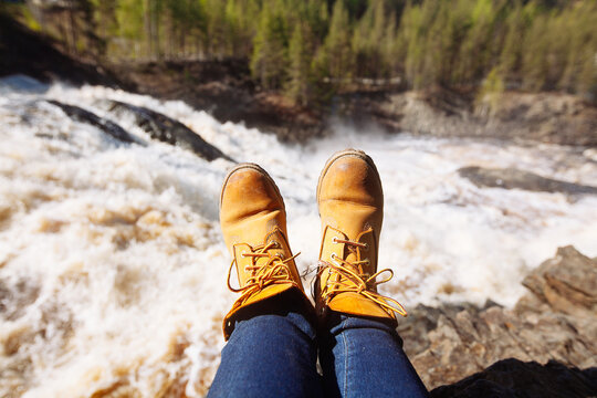 Woman's legs in blue jeans and light  brown boots sitting on the edge near waterfall