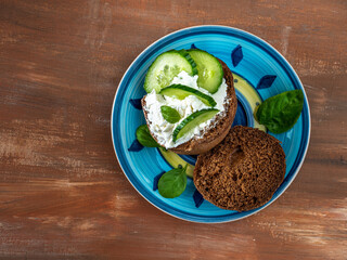 Sandwich on a rye bun with soft cheese and cucumber with basil leaves on a colored plate
