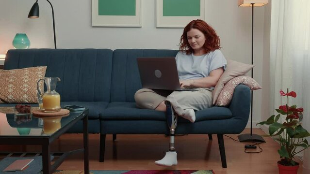 Happy attractive disabled female blogger with futuristic bionic leg working online using laptop at home looking away.