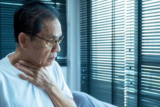senior asian man coughing wearing glasses, after waking up in the morning, in the bedroom, wearing a white shirt, hand on neck look out the window with unhealthy