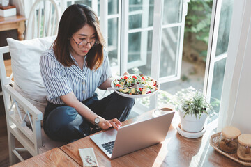 Asian woman holding a salad and work using laptop on the table in cafe. Asian woman eating salad...
