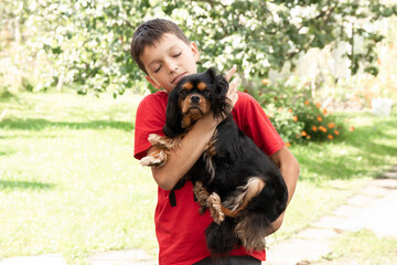 Young calm, close eyes boy in red T-shirt holding arms stroking embracing puppy dog. Relaxation, resting in countryside.