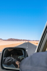Long Sahara road trip in empty reg and sandy desert, rocky mountains seen from the rear side a moving car, tuareg driver non recognisable is visible in the side mirror his arm outside the 4x4 vehicle. © MSC