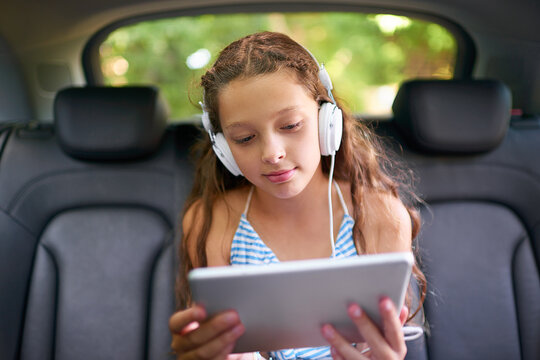 Watching her movies in the backseat. Shot of a young girl sitting in a car backseat wearing headphones and using a digital tablet.