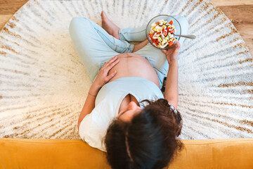 A pregnant woman holds a bowl of vegetable salad sitting on the carpet, top view, the concept of...