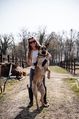 Beautiful, smiling young woman hugging big dog standing on its hind legs, having great fun together. Countryside in springtime.