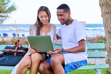 Young couple looking at laptop screen, sitting on bench on the beach