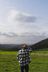 Fototapeta na wymiar Chico grande con camisa y sombrero frente a paisaje montañoso levantando los brazos y agarrando sombrero en señal de libertad
