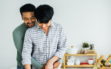 Gay LGBT sweet Asian couple wearing pajamas, smiling, hugging with happiness and love after wake up while making breakfast with healthy fruits in kitchen at home in morning. Lifestyle Concept.