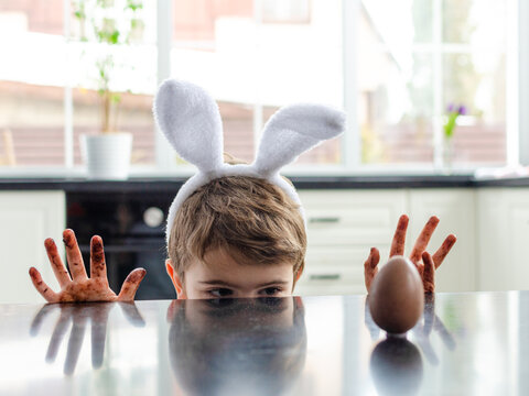 A boy with rabbit ears on his head looks at a chocolate egg