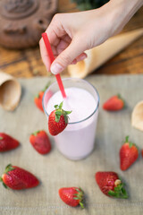 Hand holds a straw of a strawberry milkshake. Smoothie. Strawberries and ice cream cones on the table. Natural background.