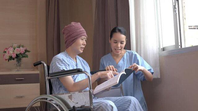 An Asian Male Cancer Patient In A Head Cover Sitting On A Wheelchair Reading A Book In Hand Together With A Smiling Asian Female Nurse Near A Window In The Patient Room During Chemotherapy In Hospital