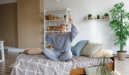 a young guy in gray pajamas is dancing sitting on the bed with his hands up.