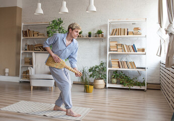 a young happy man sings and dances with a broom to clean a room at home.