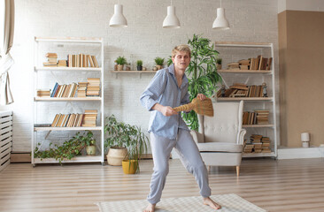 a young happy man sings and dances with a broom to clean a room at home.