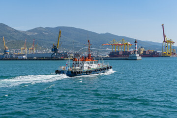 Obraz premium Blue pilot tug is moving along bay. In background are the piers, port cranes and mountains of Tsemess Bay. Close-up. Novorossiysk commercial sea port. Novorossiysk, Russia - September 15, 2021