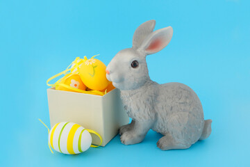 A beautiful gray rabbit toy next to a white wooden box, with yellow eggs on a blue background. Easter concept, celebration, decoration.
