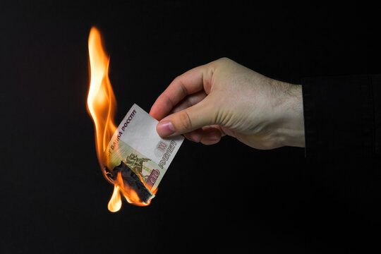 Close-up photo, a man's hand holds an almost burnt banknote of 100 rubles on a dark background.