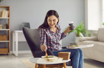 Happy woman eating tasty takeaway lunch at home. Beautiful young girl sitting in armchair in living...