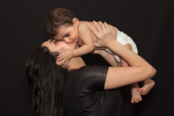 A young mother holds her little son over her head and kisses him. Isolated portrait on black background.