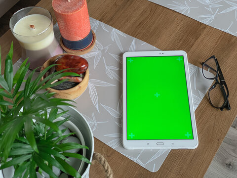 White tablet with green screen laying on wooden table next to candle, natural stones and green plant
