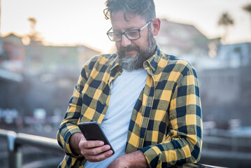Mature bearded man use mobile phone with internet wireless connection wearing glasses. Adult male people chatting with smartphone outside. Sunlight in background. Leisure activity